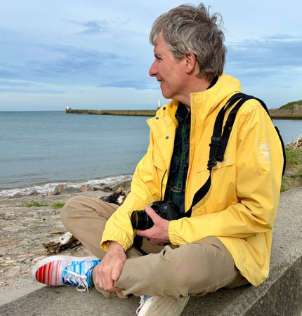 Photo of Prof Colin Moffat at the seaside sitting on the beach with a camera in hand
