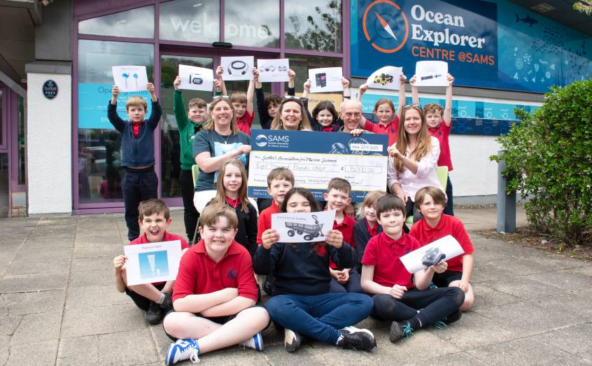 Robert Kincaid presents a cheque for £8,000 to SAMS outreach staff as children from Dunbeg Primary School pupils, who will be among the first to use the equipment, look on.